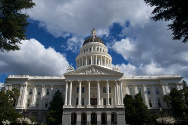 California State Capital Building from a distance bordered by trees and a blue sky with grey and white clouds